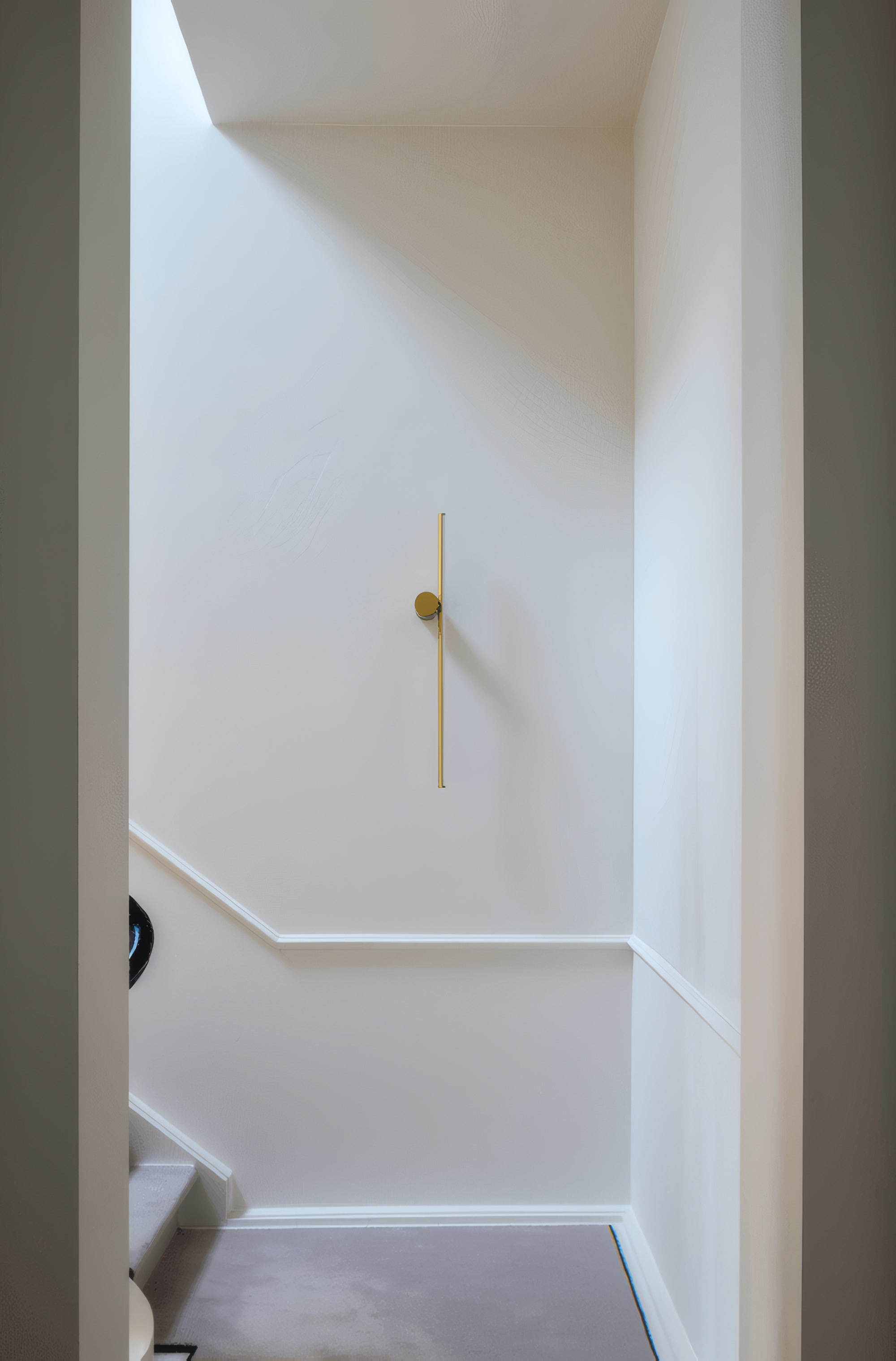 Cimaise blanche suivant la montée d’un escalier, sous une applique dorée (photo Orac Decor)